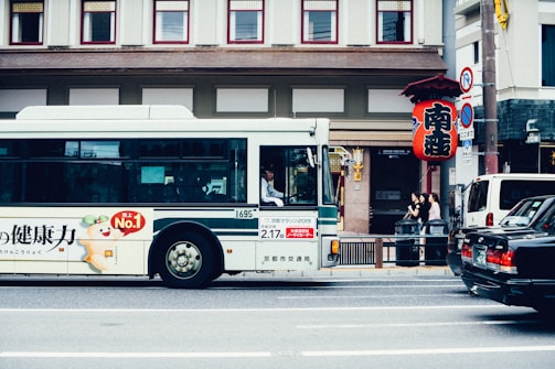 A white city bus with advertising on its side is parked on a street, with a driver visible through the window. In the background, there's a traditional-style building and a large red Japanese lantern hanging beside the sidewalk. Three people are standing near a fence on the sidewalk.