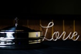 A metallic object with a small cross on top, possibly a religious item, is placed on a dark surface. To the right, the word 'love' is spelled out in a cursive style using a rope-like material. The background is dimly lit, giving a warm yet muted atmosphere.
