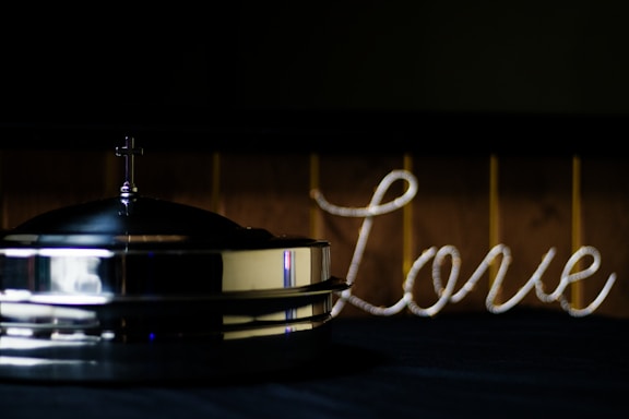 A metallic object with a small cross on top, possibly a religious item, is placed on a dark surface. To the right, the word 'love' is spelled out in a cursive style using a rope-like material. The background is dimly lit, giving a warm yet muted atmosphere.