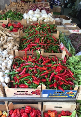 A vibrant display of fresh vegetables arranged in wooden crates at a market. The foreground is dominated by an array of red and green chili peppers. Surrounding them are other vegetables like white onions, garlic, ginger, and green and red bell peppers, along with some leafy greens and potatoes in the back.