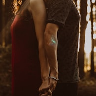 Two people stand back to back, holding hands. The scene is set in a forest with sunlight filtering through the trees, creating a warm and intimate atmosphere.