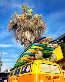A brightly colored van is parked with multiple surfboards stacked on its roof. In the background, a tall palm tree stands against a clear blue sky.
