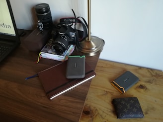 On a wooden desk, there is a digital camera with a lens cap, a stack of books, an open laptop, a metallic desk lamp, a large camera lens, and multiple wallets. One of the books is a leather-bound notebook and several hard drives are placed on top of it.