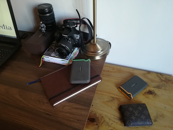 On a wooden desk, there is a digital camera with a lens cap, a stack of books, an open laptop, a metallic desk lamp, a large camera lens, and multiple wallets. One of the books is a leather-bound notebook and several hard drives are placed on top of it.