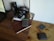 On a wooden desk, there is a digital camera with a lens cap, a stack of books, an open laptop, a metallic desk lamp, a large camera lens, and multiple wallets. One of the books is a leather-bound notebook and several hard drives are placed on top of it.