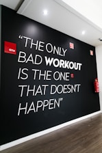 A motivational quote on a black wall in a gym setting reads 'The only bad workout is the one that doesn't happen.' The words are in large, bold white letters with 'workout' in a striking gray. Accents include red gym signs and a fire extinguisher nearby. The setting has ceiling spotlights and wooden flooring.