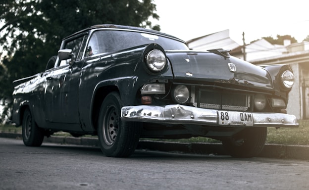 An old, vintage car is parked on the side of a street, showing signs of wear, with its dark exterior and noticeable dents on the bumper. The car is positioned against the backdrop of trees and residential buildings.
