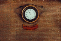 A vintage-style wall clock with a white face and black numerals is mounted against a brick wall. The clock has a wooden frame and displays a time of approximately 10:10. Below the clock, a wooden plaque with the words 'A la Graduandos Verano 2009' is attached.