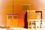 A building facade with orange corrugated metal siding featuring doors with metal grates and a large vent. A yellow wooden barricade stands in front of the doors, casting a shadow on the ground.