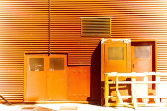 A building facade with orange corrugated metal siding featuring doors with metal grates and a large vent. A yellow wooden barricade stands in front of the doors, casting a shadow on the ground.