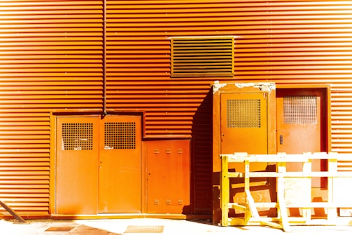 A building facade with orange corrugated metal siding featuring doors with metal grates and a large vent. A yellow wooden barricade stands in front of the doors, casting a shadow on the ground.