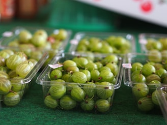 Several plastic containers filled with green gooseberries arranged on a market stall covered in green fabric. The fruits are fresh and have faint striped patterns on the skin.