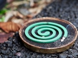 A green spiral mosquito coil is placed on a round metal holder. The background features a mix of rough, textured ground and blurred natural elements, suggesting an outdoor setting.