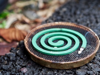 A green spiral mosquito coil is placed on a round metal holder. The background features a mix of rough, textured ground and blurred natural elements, suggesting an outdoor setting.