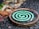 A green spiral mosquito coil is placed on a round metal holder. The background features a mix of rough, textured ground and blurred natural elements, suggesting an outdoor setting.
