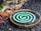 A green spiral mosquito coil is placed on a round metal holder. The background features a mix of rough, textured ground and blurred natural elements, suggesting an outdoor setting.