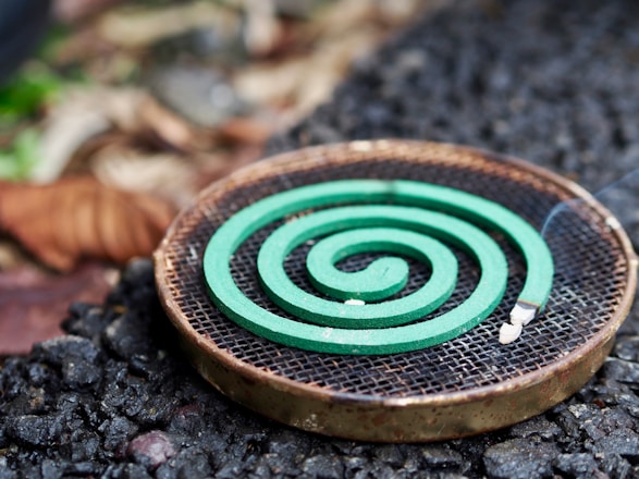 A green spiral mosquito coil is placed on a round metal holder. The background features a mix of rough, textured ground and blurred natural elements, suggesting an outdoor setting.