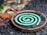 A green spiral mosquito coil is placed on a round metal holder. The background features a mix of rough, textured ground and blurred natural elements, suggesting an outdoor setting.