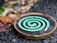 A green spiral mosquito coil is placed on a round metal holder. The background features a mix of rough, textured ground and blurred natural elements, suggesting an outdoor setting.