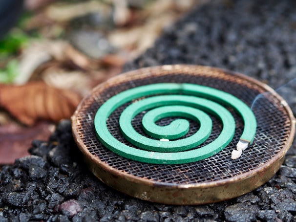 A green spiral mosquito coil is placed on a round metal holder. The background features a mix of rough, textured ground and blurred natural elements, suggesting an outdoor setting.