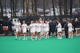 A group of lacrosse players wearing white uniforms are gathered on a green field. They are holding lacrosse sticks and appear to be preparing for or concluding a game. Some players are wearing headbands, and there are people in black jackets nearby, possibly coaches or team staff. The background features leafless trees and parked cars.