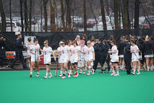 A group of lacrosse players wearing white uniforms are gathered on a green field. They are holding lacrosse sticks and appear to be preparing for or concluding a game. Some players are wearing headbands, and there are people in black jackets nearby, possibly coaches or team staff. The background features leafless trees and parked cars.