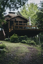 A rustic wooden cabin with a spacious deck surrounded by lush greenery and tall trees. The cabin features a sloped roof with shingle tiles and a chimney. On the deck, there is a folded umbrella and some chairs, suggesting a space for relaxation. The foreground is a pathway leading uphill to the cabin, with patches of grass and small shrubs along the way.
