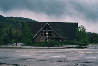 An abandoned A-frame building situated in a rural area surrounded by overgrown vegetation. The sky is overcast, creating a somber atmosphere. The building appears weathered and neglected, with partially boarded windows and a generally aged appearance. The cracked and wet pavement in the foreground adds to the desolate environment.