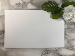 A blank white sheet of paper lies on a marble-textured surface. In the top right corner, there is a white artificial rose with two large green leaves attached to its stem.