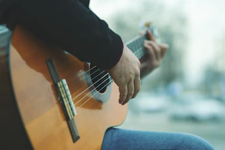 A person is playing an acoustic guitar, focusing on the hands strumming the strings. The guitar is made of wood and the background appears to be blurred, indicating an outdoor setting.