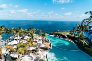A luxurious resort setting overlooking a vast ocean. Palm trees line the area, with white sun umbrellas and loungers arranged next to a curvaceous swimming pool. The water's azure hue complements the clear sky, creating a serene and inviting atmosphere. A few people are enjoying the view and poolside amenities.