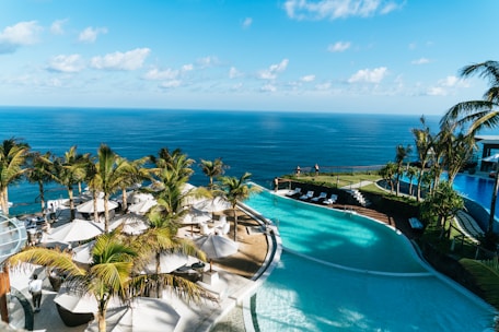 A luxurious resort setting overlooking a vast ocean. Palm trees line the area, with white sun umbrellas and loungers arranged next to a curvaceous swimming pool. The water's azure hue complements the clear sky, creating a serene and inviting atmosphere. A few people are enjoying the view and poolside amenities.