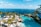A luxurious resort setting overlooking a vast ocean. Palm trees line the area, with white sun umbrellas and loungers arranged next to a curvaceous swimming pool. The water's azure hue complements the clear sky, creating a serene and inviting atmosphere. A few people are enjoying the view and poolside amenities.