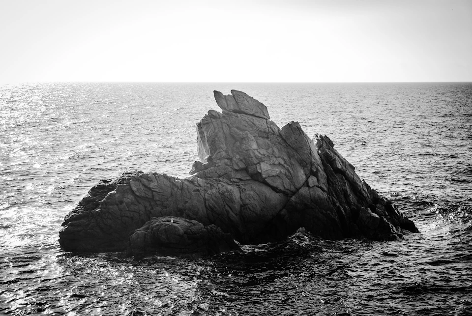 The image depicts a rocky outcropping jutting into the ocean, with the vast expanse of water surrounding it. The rock formation is composed of large boulders and smaller rocks, creating a rugged and natural landscape. The photograph is taken from a distance, allowing for a comprehensive view of the scene. The black and white color scheme adds a timeless quality to the image, emphasizing the textures and shapes within the rocky outcropping.