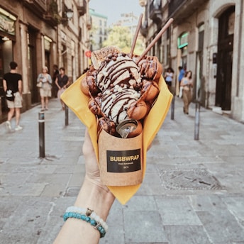 A hand holds a dessert wrapped in a brown paper cone with the label 'Bubbwrap'. The dessert consists of a bubble waffle topped with scoops of ice cream, chocolate drizzle, chocolate balls, a toasted marshmallow, and two wafer sticks. The scene is set in an urban street with people walking in the background.