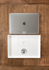 A closed silver laptop with a prominent logo sits on a wooden table. Below it, an open, empty laptop box made of white material showcases a minimalist design. The box's interior includes a space for accessories, indicating it is newly opened or ready for packaging.