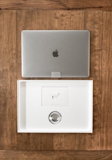 A closed silver laptop with a prominent logo sits on a wooden table. Below it, an open, empty laptop box made of white material showcases a minimalist design. The box's interior includes a space for accessories, indicating it is newly opened or ready for packaging.