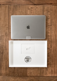 A closed silver laptop with a prominent logo sits on a wooden table. Below it, an open, empty laptop box made of white material showcases a minimalist design. The box's interior includes a space for accessories, indicating it is newly opened or ready for packaging.