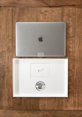 A closed silver laptop with a prominent logo sits on a wooden table. Below it, an open, empty laptop box made of white material showcases a minimalist design. The box's interior includes a space for accessories, indicating it is newly opened or ready for packaging.
