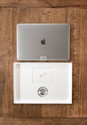 A closed silver laptop with a prominent logo sits on a wooden table. Below it, an open, empty laptop box made of white material showcases a minimalist design. The box's interior includes a space for accessories, indicating it is newly opened or ready for packaging.