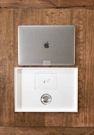 A closed silver laptop with a prominent logo sits on a wooden table. Below it, an open, empty laptop box made of white material showcases a minimalist design. The box's interior includes a space for accessories, indicating it is newly opened or ready for packaging.