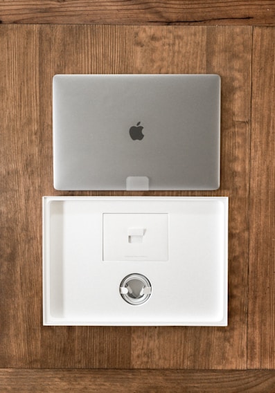 A closed silver laptop with a prominent logo sits on a wooden table. Below it, an open, empty laptop box made of white material showcases a minimalist design. The box's interior includes a space for accessories, indicating it is newly opened or ready for packaging.