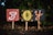 Three hand-painted signs on sticks are placed in front of lush green foliage. Each sign has a different colored background and displays a painted letter, together forming the word 'JOY'. The first sign is red with a white and multicolored 'J', the middle sign is black with a yellow 'O', and the last sign is black with an orange 'Y'. Fallen leaves and grass are visible on the ground.