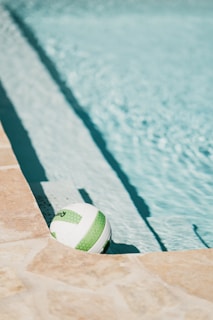 A volleyball with green and white patterns rests against the edge of a swimming pool. The pool water is a vibrant light blue, and the sun casts shadows on the stone deck that surrounds the pool.