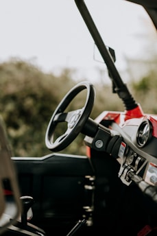 A close-up view of the interior of a vehicle focusing on the steering wheel and dashboard. The vehicle appears to be parked in an outdoor setting, with a blurred, natural background visible through the windshield. The dashboard is equipped with various controls and gauges.