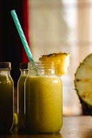 A large glass jar filled with a yellow-green smoothie is topped with a slice of pineapple. A blue and white striped straw sticks out of the jar. Two similar bottles are partially visible in the background, and a halved pineapple sits to the side. The scene is warmly lit, suggesting a cozy atmosphere.