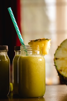 A large glass jar filled with a yellow-green smoothie is topped with a slice of pineapple. A blue and white striped straw sticks out of the jar. Two similar bottles are partially visible in the background, and a halved pineapple sits to the side. The scene is warmly lit, suggesting a cozy atmosphere.