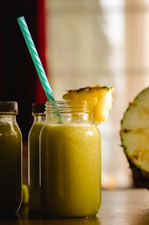 A large glass jar filled with a yellow-green smoothie is topped with a slice of pineapple. A blue and white striped straw sticks out of the jar. Two similar bottles are partially visible in the background, and a halved pineapple sits to the side. The scene is warmly lit, suggesting a cozy atmosphere.