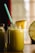 A large glass jar filled with a yellow-green smoothie is topped with a slice of pineapple. A blue and white striped straw sticks out of the jar. Two similar bottles are partially visible in the background, and a halved pineapple sits to the side. The scene is warmly lit, suggesting a cozy atmosphere.