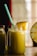 A large glass jar filled with a yellow-green smoothie is topped with a slice of pineapple. A blue and white striped straw sticks out of the jar. Two similar bottles are partially visible in the background, and a halved pineapple sits to the side. The scene is warmly lit, suggesting a cozy atmosphere.